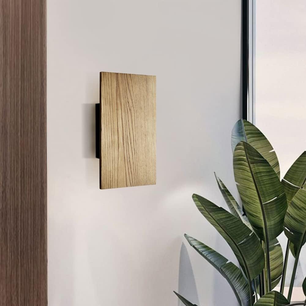 Wooden wall-mounted light fixture on a white wall with a plant in the foreground
